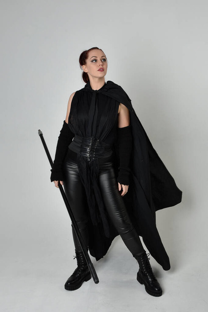 Retrato de comprimento total de modelo feminino ruiva bonita vestindo traje de couro scifi futurista preto, manto preto segurando uma arma de lança. Posição de pé dinâmica no fundo do estúdio branco. - Foto, Imagem