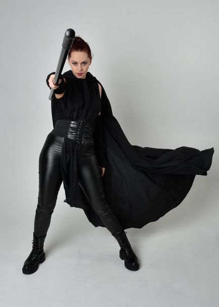 Teljes hosszúságú portré csinos vörös hajú női modellről fekete futurisztikus scifi bőr jelmezben, fekete köpenyben, kezében egy lándzsa fegyverrel. Dinamikus álló póz fehér stúdió háttér. - Fotó, kép