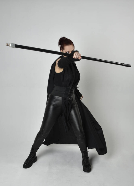 Portrait pleine longueur de jolie rousse modèle féminin portant costume futuriste noir en cuir scifi, manteau noir tenant une arme de lance. pose debout dynamique sur fond de studio blanc. - Photo, image