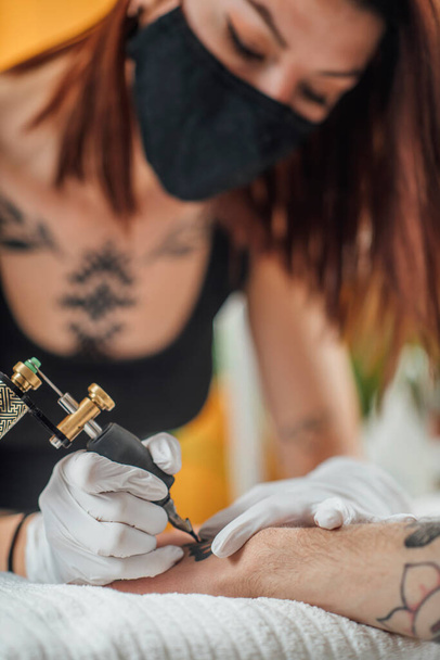 Segurança na tatuagem durante a crise do coronavírus. Tatuagem artista tatuando cliente com luvas e máscara facial em um estúdio de tatuagem durante a pandemia de COVID-19 - Foto, Imagem