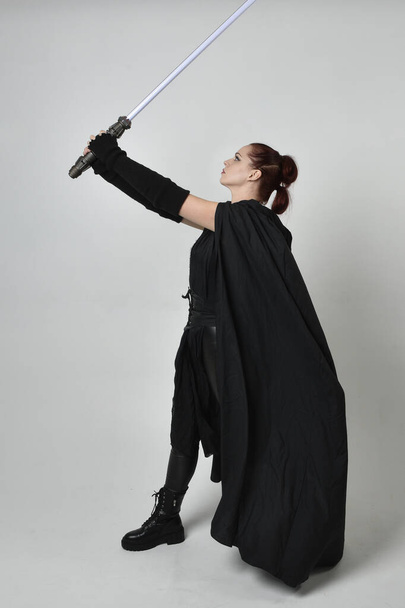 Портрет красивої рудої жіночої моделі в чорному футуристичному шкіряному плащі, що тримає зброю Lightaber. Динамічна стояча поза на білому фоні студії
. - Фото, зображення