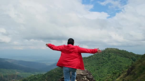 Νεαρή Ασιάτισσα πεζοπόρος με κόκκινο αδιάβροχο να στέκεται στην κορυφή του βουνού με σηκωμένα χέρια και να απολαμβάνει τη θέα. έννοια ταξίδι και επιτυχία - Πλάνα, βίντεο