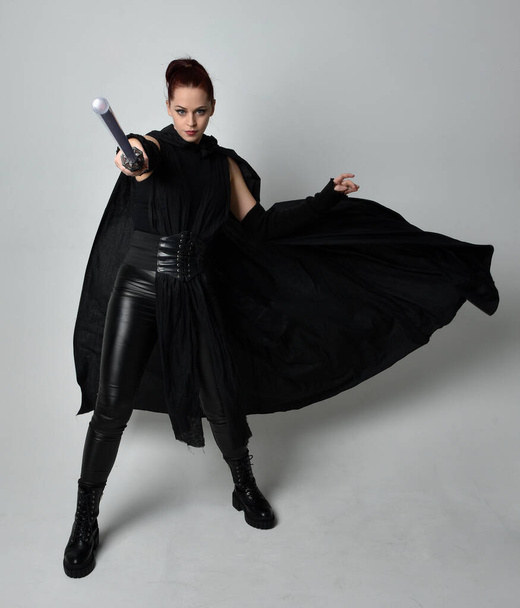 Full length portrait of pretty redhead female model wearing black futuristic scifi leather cloak costume, holding a lightsaber weapon. Dynamic standing pose on white studio background. - Fotoğraf, Görsel