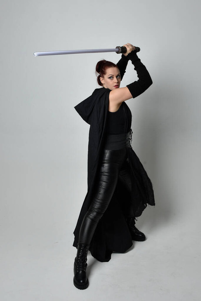 Ολο το μήκος πορτρέτο της όμορφης κοκκινομάλλας γυναίκας μοντέλο φορώντας μαύρη φουτουριστική scifi δερμάτινη στολή μανδύα, κρατώντας ένα φωτόσπαθο όπλο. Δυναμική στάση σε λευκό φόντο στούντιο. - Φωτογραφία, εικόνα