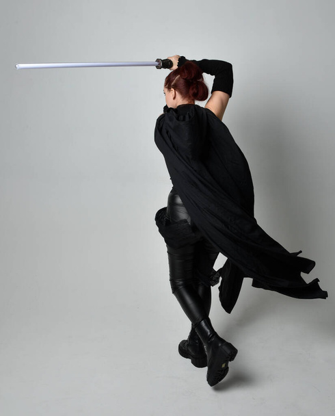 Portrait pleine longueur de jolie rousse modèle féminin portant noir futuriste manteau en cuir scifi costume, tenant une arme sabre laser. pose debout dynamique sur fond de studio blanc. - Photo, image