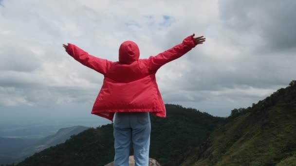 Jeune randonneuse asiatique vêtue d'un imperméable rouge debout sur le sommet de la montagne avec les bras levés et profitant de la vue. voyage et concept de succès - Séquence, vidéo