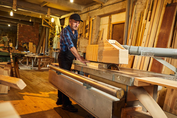 Wood cutting table with electric circular saw.  Senior Professional carpenter in uniform cutting wooden board at sawmill carpentry manufacturing . Sawing machine - Φωτογραφία, εικόνα