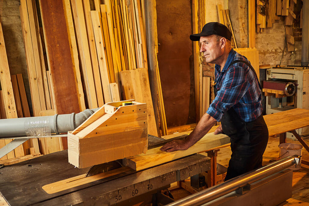 Τραπέζι κοπής ξύλου με ηλεκτρικό δισκοπρίονο. Ανώτερος Επαγγελματίας ξυλουργός σε ομοιόμορφη κοπή ξύλινων σανίδων σε ξυλουργείο ξυλουργικής. Μηχανήματα πριονίσματος - Φωτογραφία, εικόνα