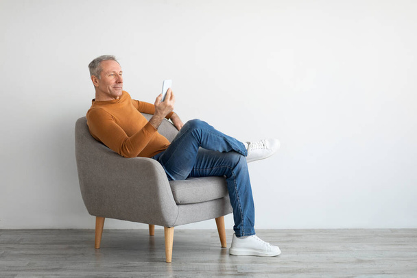 Portrait of mature man using smartphone sitting on armchair - Foto, Imagen