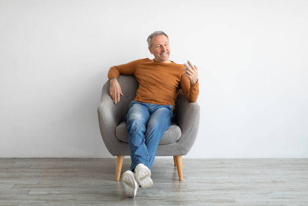 Portrét usmívajícího se dospělého muže pomocí smartphonu sedícího na křesle - Fotografie, Obrázek