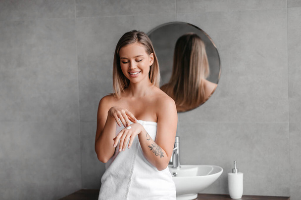 Onnellinen nainen, joka käyttää kosteuttavaa voidetta käsiin, hankaa ravitsevaa vartalovoidetta käsivarsiin, istuu kylpyhuoneessa - Valokuva, kuva