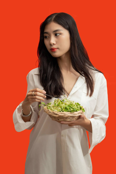 Vegetarian concept, Υγιεινή γυναίκα τρώει φύτρες ηλίανθου που στέκονται απομονωμένες σε πορτοκαλί φόντο. - Φωτογραφία, εικόνα