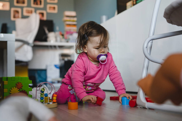 Uma pequena menina caucasiana de dez meses de idade brincando sozinha no espaço de cópia em casa usando tijolos e brinquedos vestindo rosa no chão no dia - Foto, Imagem