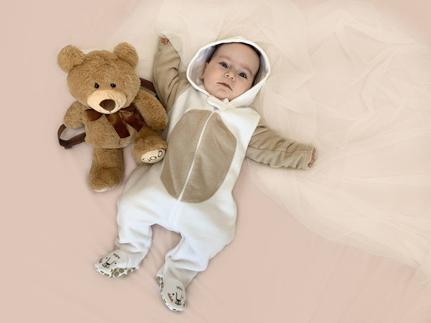 Vauva ja karhu. Kaunis hymyilevä lapsi makaa sängyllä lempipehmolelunsa vieressä. Vaakasuora kuva, ylhäältä, pastelliväri vuodevaatteet. Kopioi Käsite: lapsuus, lasten uni - Valokuva, kuva