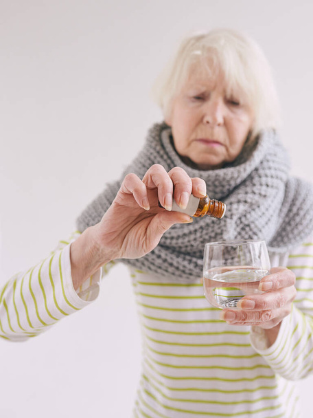 senior ill woman in scarf freezing cold put drops in glass of water on white background. Health care, crisis, oldness concept - Foto, Imagem