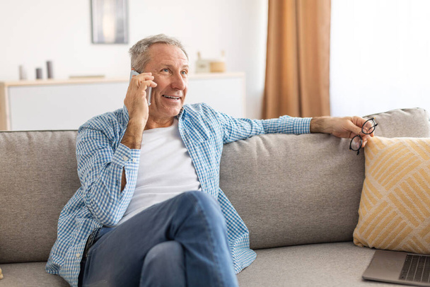 Веселый взрослый мужчина разговаривает по телефону, сидя на диване - Фото, изображение
