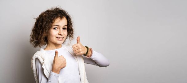 One small caucasian girl ten years old with curly hair front view portrait close up standing in front of white background looking to the camera holding thumbs up copy space smiling happy and joy - Foto, Bild