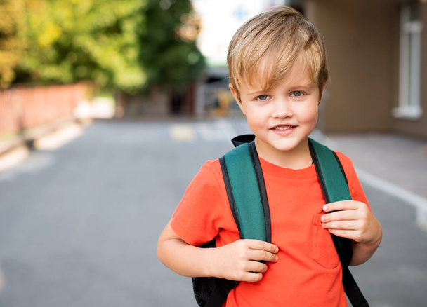 Χαμογελαστό αγόρι με κόκκινο μπλουζάκι και σακίδιο. Στέκεται στο δρόμο. - Φωτογραφία, εικόνα