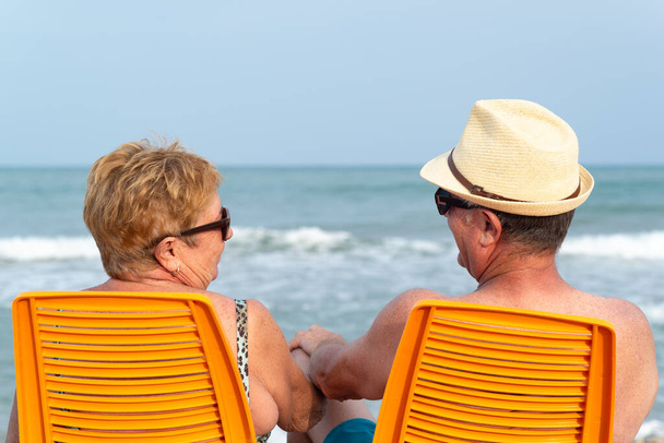Birbirlerine aşık yaşlı bir çiftin kumsalda otururken el ele tutuşup el ele tutuşması. Rahatlamış yaz tatili. - Fotoğraf, Görsel