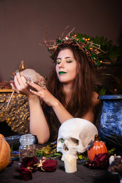 Portrait of a witch with tousled hair and long nails with a wreath of dry branches on her head, dressed in a green dress, holding a white rat. - Foto, Imagem