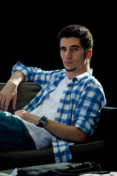 Elegant young man with a wristwatch leaning on a bench while looking at the camera, with a dark background behind it - Photo, Image