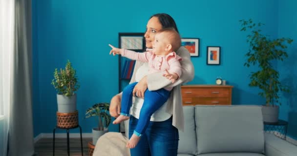 Llena de fuerza madre sostiene al niño en brazos frente a la ventana, señala con el dedo en frente - Imágenes, Vídeo