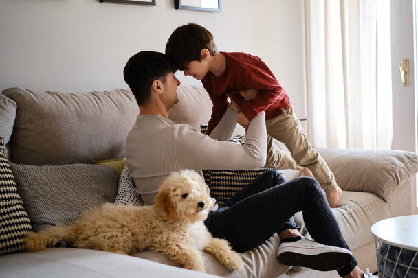 Πατέρας και γιος διασκεδάζουν μαζί ενώ χαλαρώνουν στο σπίτι με το σκύλο τους. - Φωτογραφία, εικόνα