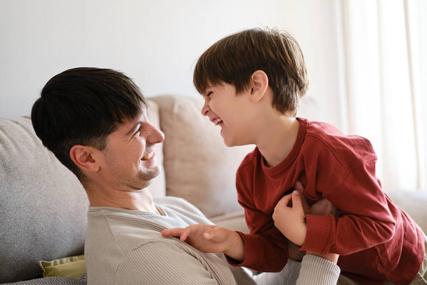 Glücklicher Vater und Sohn schauen einander an und lachen, während sie zu Hause zusammen genießen und spielen. - Foto, Bild