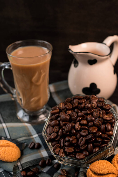 Des grains de café noirs dans un pot en verre à côté de la tasse de cappuccino et de la cruche de lait - Photo, image