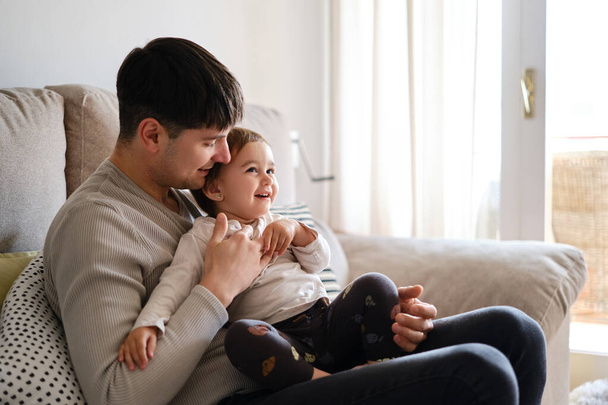 Homem aproveitando o tempo com sua filhinha enquanto relaxa juntos em um sofá em casa. - Foto, Imagem