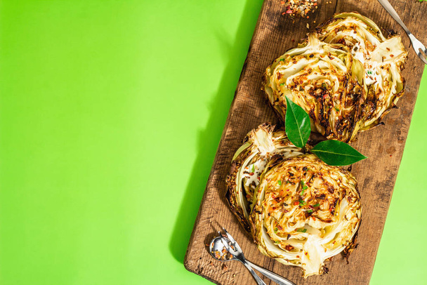 Σπιτικές vegan μπριζόλες λάχανο με βότανα και μπαχαρικά. Υγιεινά τρόφιμα έτοιμα για κατανάλωση σε μια ξύλινη επιφάνεια κοπής. Σύγχρονο σκληρό φως, σκούρα σκιά, φωτεινό πράσινο φόντο, κορυφαία άποψη - Φωτογραφία, εικόνα