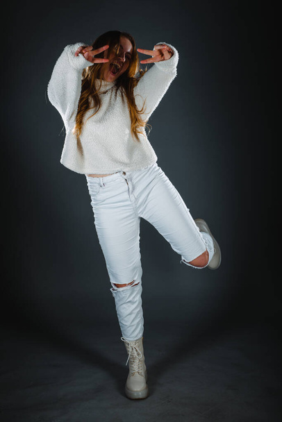 Junge Fashionista in weißen Jeans und mit nacktem Bauch posiert vor dunkler Wand - Foto, Bild