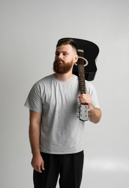 Handsome brunette bearded man musician standing and holding a acoustic guitar on a shoulder on a white background studio. - Photo, Image