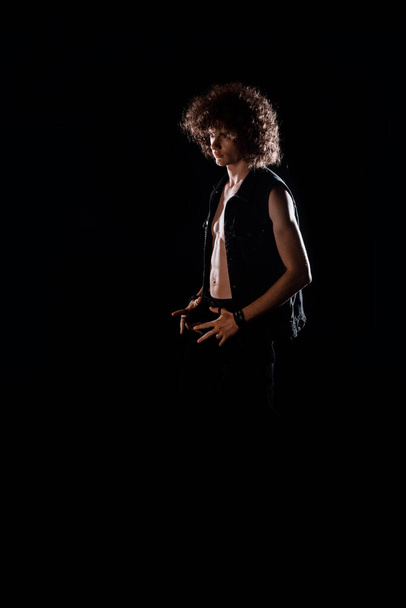 Przystojny młody chłopiec w hipsterskiej kamizelce dżinsów podczas pozowania na ciemne tło - Zdjęcie, obraz