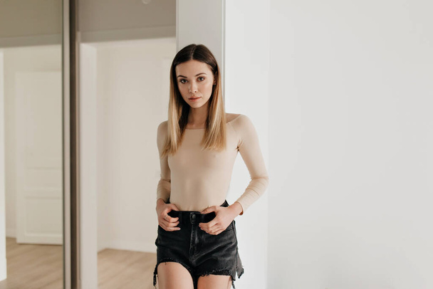 Pretty charming girl wearing beige body and black shorts is posing at camera in modern stylish apartment. Lifestyle, leisure concept. High quality photo - Fotó, kép