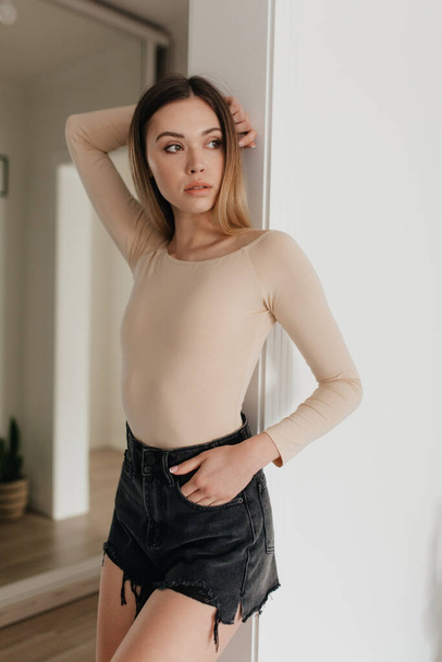 Relaxed cheerful blond woman in stylish beige pullover and shorts smiles sincerely and looking aside. Happy pretty girl stands over white wall. High quality photo - Foto, afbeelding