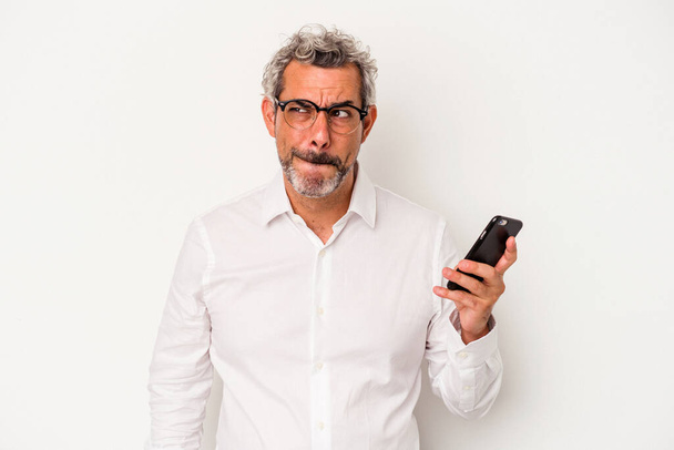 Mittelalter kaukasischen Geschäftsmann hält ein Handy isoliert auf weißem Hintergrund verwirrt, fühlt sich zweifelhaft und unsicher. - Foto, Bild