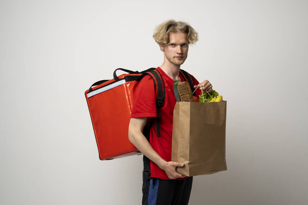 Food delivery service. Portrait of pleased delivery man in red uniform smiling while carrying paper bag with food products isolated over white background. - Fotografie, Obrázek