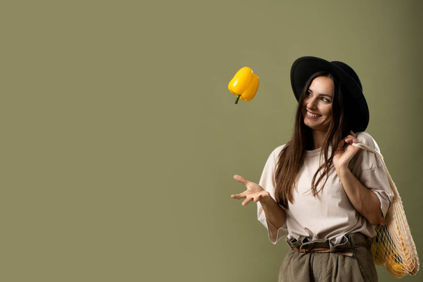 Eco friendly smiling woman in beige t-shirt and black hat throw up a yellow pepper in a air by one hand and holding reusable cotton eco bags full of groceries on a shoulder. - Photo, Image