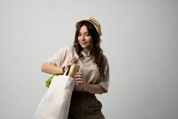 Smiling young woman in beige t-shirt and a hst with a mesh eco bag full of vegetables and on a white studio background. Sustainable lifestyle. Eco friendly concept. Zero waste. - Фото, зображення