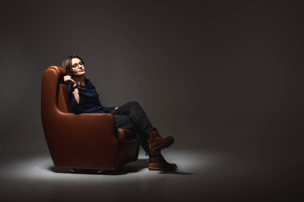 Mulher bonita, jovem, emocional, cansada e frustrada sentada em uma cadeira de couro marrom em uma sala isolada, um feixe de luz ilumina. Chave baixa. Dias difíceis - Foto, Imagem