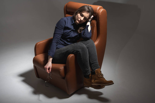 Belle, jeune, émotionnelle, fatiguée et frustrée femme assise sur une chaise en cuir marron dans une pièce isolée isolée, un faisceau de lumière éclaire. Très discret. Des jours durs - Photo, image