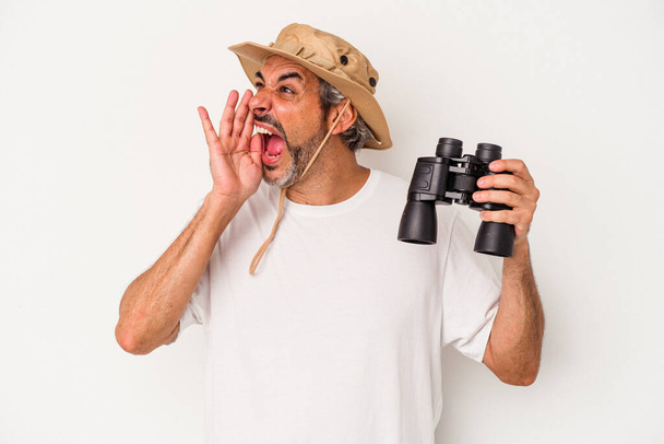 Middelbare leeftijd blanke man met verrekijker geïsoleerd op witte achtergrond schreeuwen en vasthouden palm in de buurt van geopende mond. - Foto, afbeelding