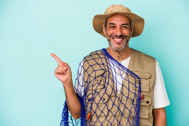 Pescador caucásico de mediana edad sosteniendo red aislada sobre fondo azul sonriendo y señalando a un lado, mostrando algo en espacio en blanco. - Foto, Imagen