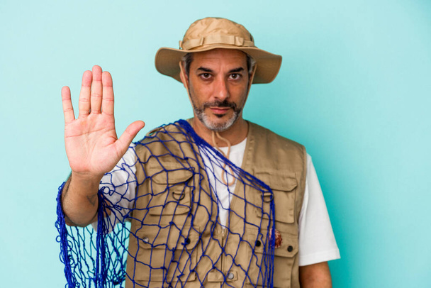 Pescador caucásico de mediana edad sosteniendo red aislada sobre fondo azul de pie con la mano extendida mostrando señal de stop, lo que le impide. - Foto, Imagen