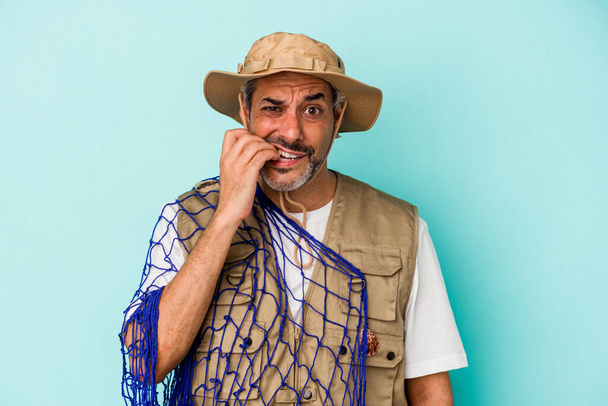 Pescador caucásico de mediana edad sosteniendo red aislada sobre fondo azul mordiendo las uñas, nervioso y muy ansioso. - Foto, Imagen