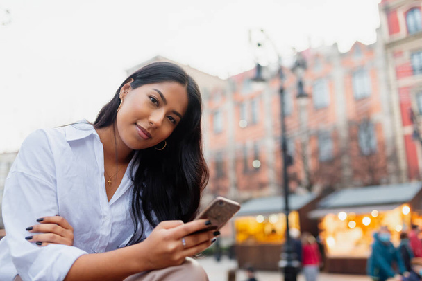portrait de jeune femme latine regardant la caméra et tenant son téléphone portable dans une rue de la ville. - Photo, image