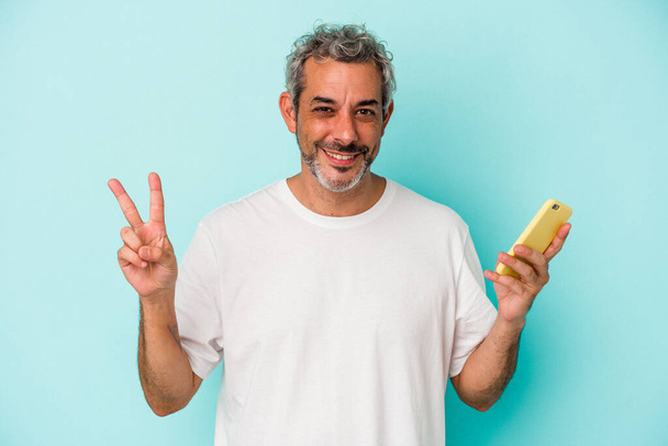 Hombre caucásico de mediana edad sosteniendo un teléfono móvil aislado sobre fondo azul mostrando el número dos con los dedos. - Foto, Imagen