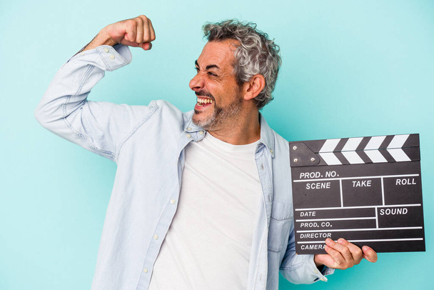 Middle age caucasian man holding a clapperboard isolated on blue background  raising fist after a victory, winner concept. - Foto, Imagen