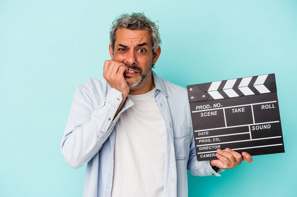 Middle age caucasian man holding a clapperboard isolated on blue background  biting fingernails, nervous and very anxious. - Foto, Imagen